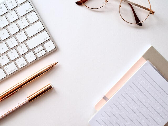 A desk with a Mac keyboard, pair of glasses, gold pens and pink and white legal pad of paper.